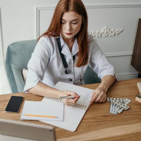 A female doctor writes notes at her desk in a clinical setting, focusing on healthcare tasks.