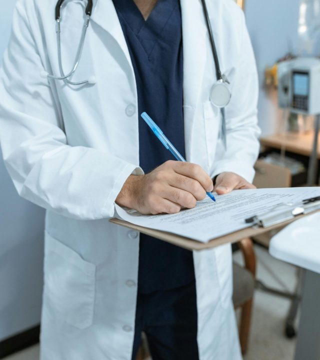 pexels-photo-6129043-6129043 A doctor in a lab coat writing notes in a hospital setting, using a clipboard.